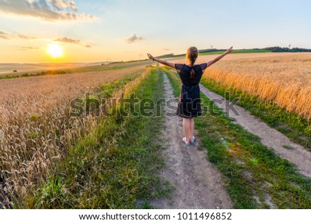 Young girl standing on dirt gravel road in wheat field at sunset raising hands