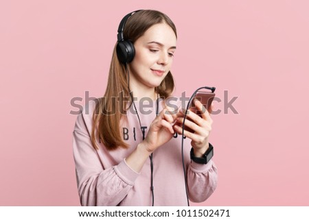 Studio shot of pleasant looking femal reads news online on cellphone or watches video in headphones, uses free internet connection, isolated over pink background. Young girl messages with friends