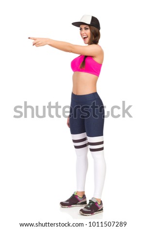 Young woman in pink sports bra, leggings and cap is looking away, laughing and pointing. Full length studio shot isolated on white.