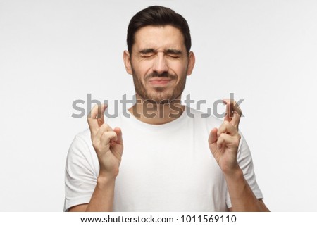 Closeup portrait of handsome caucasian man with closed eyes in white tshirt isolated on gray background, crossing fingers for luck