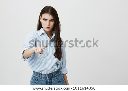 Studio portrait of young european woman in blue-collar shirt, expressing aggression with eyes and frowning while pointing index finger at camera, standing over gray background.