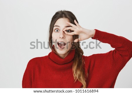 Horizontal shot of funny positive joyful girl with ponytail in red sweater with opened mouth, smiles joyfully, looks at camera through ok sign, has playful mood. Body language and face expression