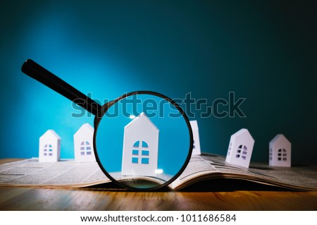 Magnifying glass in front of an open newspaper with paper houses. Concept of rent, search, purchase real estate.