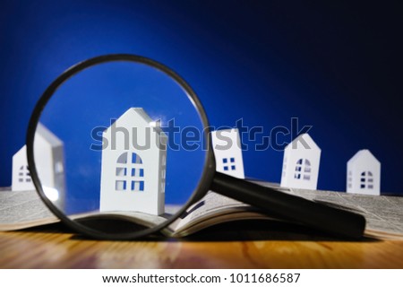 Magnifying glass in front of an open newspaper with paper houses. Concept of rent, search, purchase real estate.