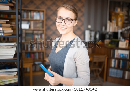 young smiling caucasian woman in glasses is standing with a book in the hands of a book rack