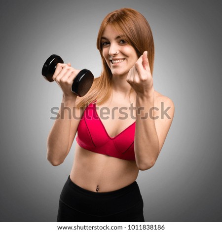 Beautiful sport woman with dumbbells making money gesture on grey background