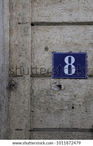 Close uo view of the number eight written in white on a blue metallic rectangle. Element fixed on an house grey stone wall in a street in France. Graphic picture. Simple sign. Geometric design. 