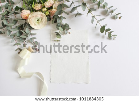 Feminine wedding, birthday desktop mock-up scene. Blank paper greeting card and bouquet of eucalyptus branches, pink roses and Persian buttercup flowers. White table background. Flat lay, top  view.