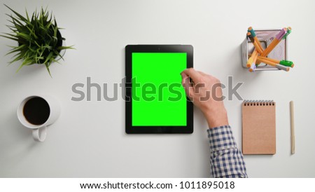A man's finger scrolling on the green touchscreen. A tablet is on the white table. View from the top. Close-up.