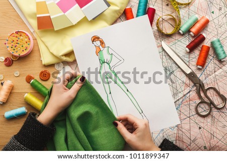 Top view of fashion designer working with material sample and hand-drawn sketch at messy table background, top view. Dressmaking, creativity and sewing workshop concept