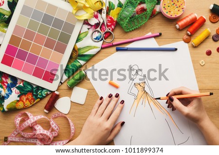 Top view on fashion designer at work. Female hands drawing clothes sketch at her creative workspace and using digital tablet with color swatches on screen, top view