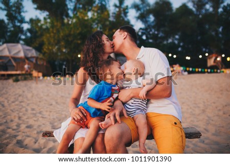 The concept of a family vacation. Young family and two sons sitting on a bench in the evening on a sandy beach. Mom and Dad kiss, the older brother kisses the younger on the lips.