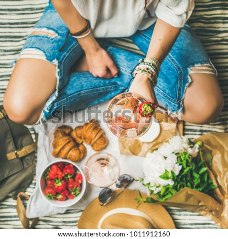 French style romantic picnic setting. Woman in jeans with glass of wine, strawberries, croissants, brie cheese, sunglasses, peony flowers on blanket, top view, square crop. Outdoor gathering concept
