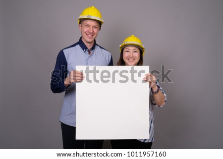 Studio shot of young handsome businessman and mature Asian businesswoman wearing hardhat against gray background