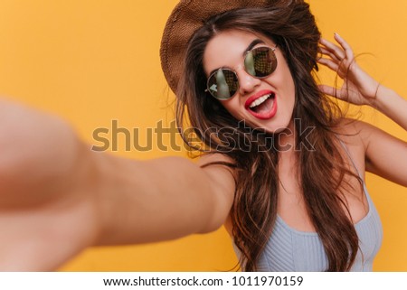 Close-up portrait of good-looking excited woman taking picture of herself. Funny girl in sunglasses making selfie on yellow background.
