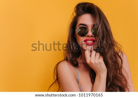 Close-up portrait of adorable brown-haired girl posing with interested smile. Indoor photo of pretty caucasian lady in glasses isolated on yellow background.