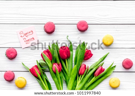 Waiting for spring. Spring is coming lettering near bouqet of red tulips and macarons on white wooden background top view space for text