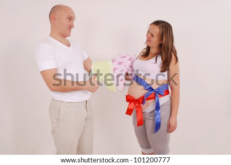 Man and pregnant woman holding baby clothes on grey background