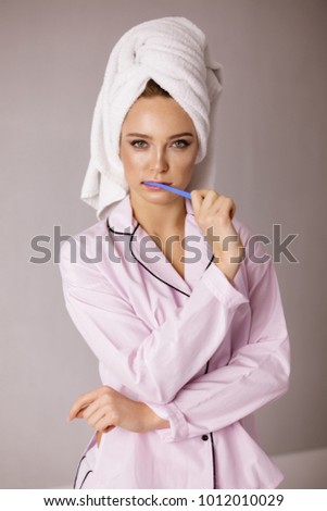 Portrait of  beautiful lady in pink sleepwear standing with towel on her head and brushing her teeth while dreamily looking in camera isolated