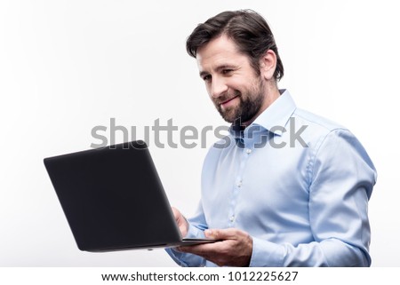 Love my job. Charming middle-aged businessman in a baby blue shirt working on his laptop and smiling pleasantly while posing against a white background