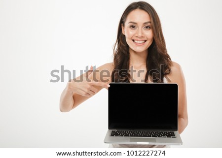 Horizontal photo of happy woman 30s smiling and demonstrating black empty screen of silver laptop on camera with finger over white wall