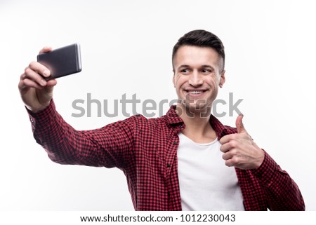 Perfect selfie. Handsome young man in a checked shirt on top of a white t-shirt taking selfies and showing thumbs up to the camera