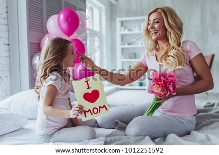I love you, mom! Attractive young woman with little cute girl are sitting on bed and spending time together at home. Mom is receiving presents from daughter on Mother's Day. Happy family concept.