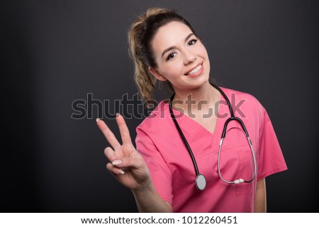 Portrait of beautiful young doctor showing peace gesture and smiling on black background with copypsace advertising area
