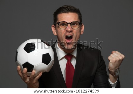 Victory! Young positive businessman in glasses and formal suit rejoices in success holding in hand ball on gray background