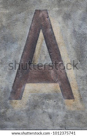 Capital letter A written on a grey ancient wall. Dark painted character on a flat vertical surface. Simple abstract picture. Symbol of the alphabet. Brown, black, grey and beige colors.  