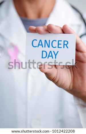 closeup of a young caucasian doctor man with a pink ribbon pinned in the flap of his white coat showing a signboard with the text cancer day written in it