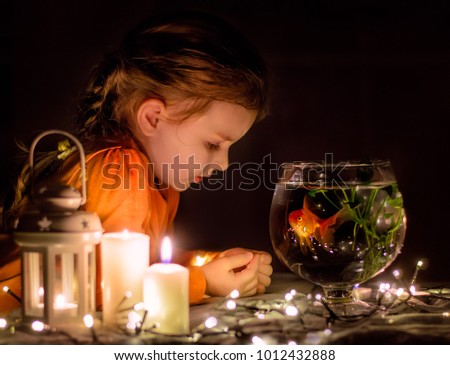 Little girl and her goldfish. Kid is making a wish to a goldfish. Child is watching  at the aquarium. Candles and garland. Magical Christmas. Christmas Eve