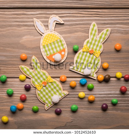 Figures of Easter bunnies lie on a wooden background decorated with round colored sweets. View from above. Flat layout. Holiday spring concept. Square frame. 