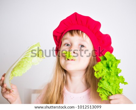 Beautiful little girl, has happy fun smiling face, big pretty eyes, long blonde hair, red hat. Cooks in  kitchen appetizing tasty vegetables. Child portrait. Creative detox concept. Close up.