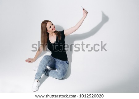 Studio full length of happy brunette woman in black t-shirt and jeans taking selfie via cell phone while sitting on the floor on white background. Isolate.