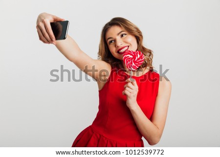 Portrait of a smiling young woman dressed in red dress holding heart shaped lollipop and taking a selfie isolated over white background