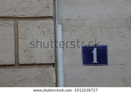 Close uo view of the number one written in white on a blue metallic rectangle. Element fixed on an house beige stone wall in a street in France. Graphic urban picture. Simple sign. Geometric design. 