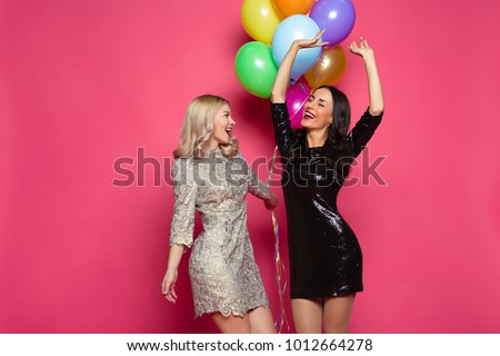 Time of celebration and fun. Two happy and cheerful beautiful girl friend in beautiful dress posing and having fun with helium balloons in hands on a pink background.