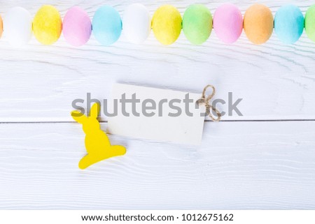 Paper blank tag decorated colorful pastel easter eggs and yellow rabbit on rustic wooden white background, top view. Happy Easter cards