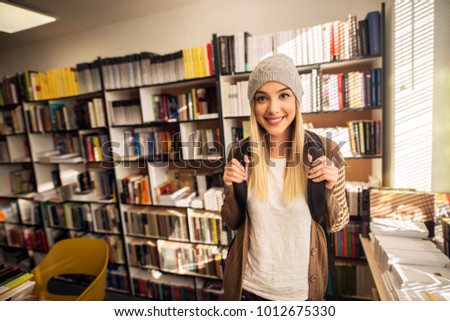 Portrait view of adorable happy attractive hardworking high school student girl standing in the sunny library or classroom and posing with backpack and hat while looking at the camera.
