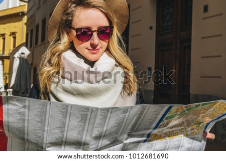 Young smiling woman tourist in hat and sunglasses stands on street of ancient European city and holds map in her hands. In background is building. Tourism, travel, vacation, trip, journey.