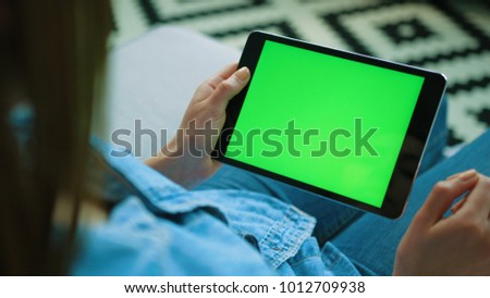 Young woman using black tablet device with green screen. Woman holding tablet, scrolling pages while sitting on the couch in the living room. Chroma key. View over shoulder. Close up