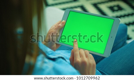 Woman using white tablet device with green screen. Woman holding tablet, scrolling pages while sitting on the couch in the living room. Chroma key. View over shoulder. Close up