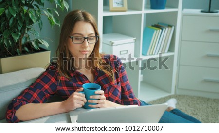 Attractive young woman wearing glasses, drinking coffee, tea, using laptop computer while sitting on the floor near couch. Portrait shot. close up