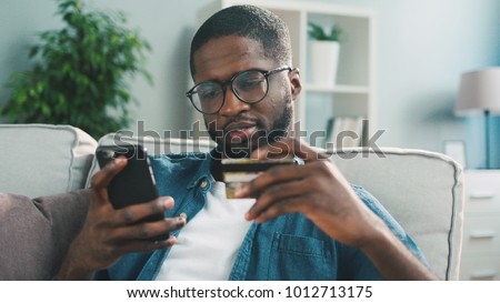 African young man in glasses shopping online with credit card using smart phone at home. Indoor.