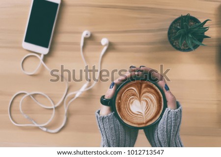 Top view female hands holding a cup of hot coffee with heart shape latte art , smartphone and cactus pot on the table.