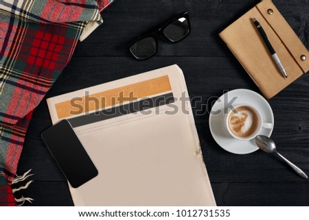 Above view of Smart phone, newspaper, scarf in a cage, glasses with notebook and cup of latte coffee on black wooden background.
