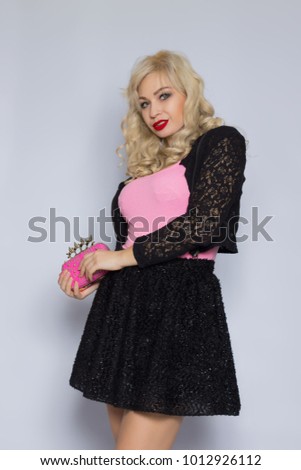 Portrait of a beautiful young blonde woman with curly hair on a gray background. The girl is dressed in a black lace suit, holds a stylish pink clutch in her hands