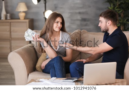 Happy woman with money and indignant man on sofa at home. Problems in relationship Royalty-Free Stock Photo #1012941076
