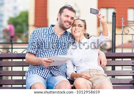 Travel Ideas and Concepts. Young Caucasian Couple in Love Sitting on Bench Outdoors While Taking Selfie Pictures.Horizontal Image Composition